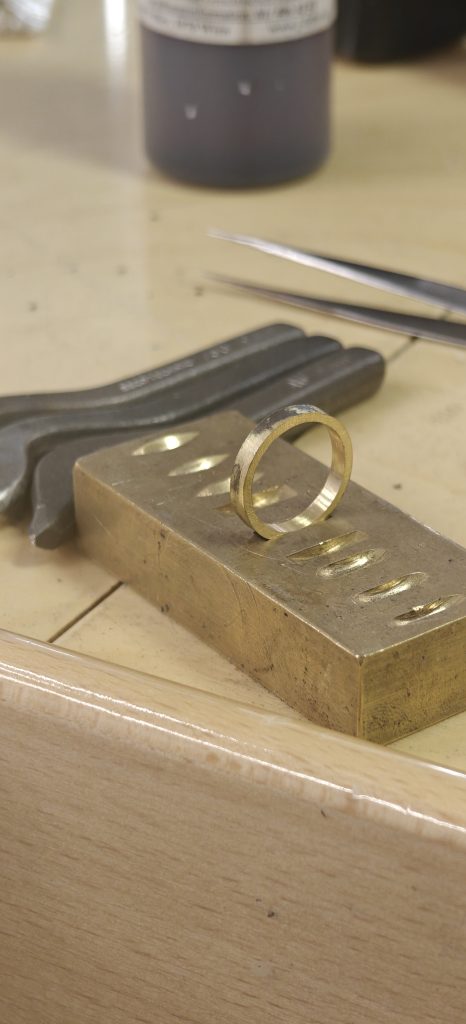 Ein Ring aus gelbem Metall (vermutlich Gold) steht auf einem Messingprägeblock. Im Hintergrund liegen Prägewerkzeuge und eine Pinzette auf dem Arbeitstisch. Der Ring zeigt sichtbare Spuren von Löt- oder Bearbeitungsarbeiten.